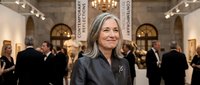Middle-aged Caucasian woman with gray hair smiling at elegant art gallery opening with classical architecture, chandelier lighting, and formal attendees.