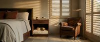 Modern bedroom interior with warm sunlight streaming through wooden shutters creating striped shadows on neutral bedding and leather armchair.