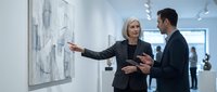 Mature blonde businesswoman explaining artwork to male colleague in modern gallery space with white walls and contemporary paintings.