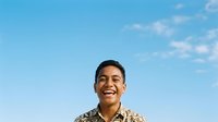 Young man with dark hair and medium brown skin tone, wearing a patterned shirt, smiling against clear blue sky. Suitable for lifestyle, happiness, youth, and portrait projects.