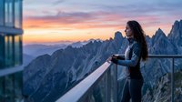 Woman with long dark hair, wearing dark athletic jacket, standing on modern mountain terrace at sunset, holding warm beverage, gazing at dramatic jagged peaks and golden sky, travel and wellness lifestyle moment.