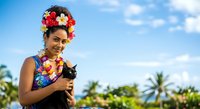 Young woman with South Asian ethnicity wearing colorful flower crown and lei, holding black cat outdoors in tropical setting with palm trees and blue sky.