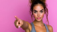 Young woman with messy bun hairstyle wearing gray sports tank top pointing finger forward against pink background, confident direct gaze and natural makeup look.