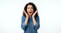Excited young Hispanic woman with curly hair in denim shirt showing surprise with hands on face against white background for marketing campaigns.
