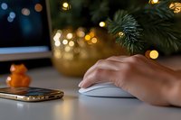 Female hand using wireless mouse near smartphone on white desk with Christmas decorations and bokeh lights, creating cozy holiday workspace atmosphere.