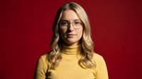 Young blonde Caucasian woman wearing glasses and yellow turtleneck sweater smiling confidently against red background for professional headshot portraits.