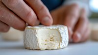 Soft creamy French cheese with white rind being cut on white surface, close up view showing texture and ripeness of gourmet dairy product.