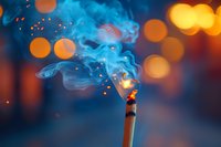 Burning incense stick with rising blue smoke against blurred warm bokeh lights background creates mystical atmospheric mood. Suitable for meditation and spa designs.