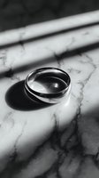 Silver wedding band on marble surface with dramatic shadows creating contrast in black and white photography.