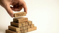 Hand placing wooden block with TRUST text on pyramid of natural wood blocks representing business foundation and reliability concepts.