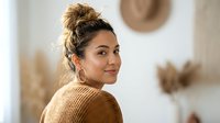 Young Hispanic woman with messy bun hairstyle wearing brown knit sweater smiling over shoulder in cozy modern home interior with neutral decor.