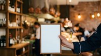Male hand in dark business sleeve holding blank white tablet menu in modern cafe with brick walls, warm lighting, and blurred customers dining in background.