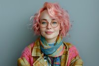 Young woman with pink wavy hair, round glasses and colorful scarf smiling at camera against blue background. Natural daylight portrait shows genuine expression.