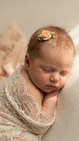 Peaceful sleeping newborn baby girl with peach flower headband wrapped in cream lace blanket for maternity photography and birth announcements.