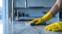 Hand in yellow rubber glove cleaning kitchen counter with sponge. Domestic housework and hygiene maintenance in modern home interior.
