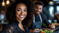 Smiling African American woman with curly hair enjoying dining experience at restaurant with blurred male server in background.