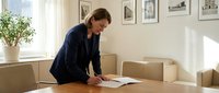 Professional businesswoman in navy suit signing documents at modern office conference table with natural lighting and framed artwork on walls.