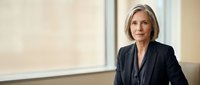 Middle-aged woman with shoulder-length blonde hair, Caucasian ethnicity, wearing dark business blazer over gray top in modern office setting with window background.