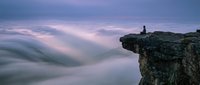 Lone figure sits on rocky cliff edge above dramatic sea of clouds at twilight, evoking solitude, freedom, and contemplation in nature.