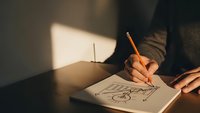 Person drawing creative sketch in notebook with pencil at wooden desk in warm natural sunlight for artistic projects and design work.