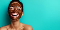 Happy woman with chocolate facial mask laughing against turquoise background, showcasing skincare routine and beauty treatment.