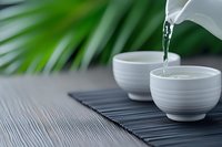 Pouring hot tea from white ceramic teapot into matching cups on bamboo mat with tropical palm leaves creating serene spa atmosphere for relaxation.