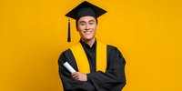 Young Asian male graduate in cap and gown holding diploma against vibrant yellow background, celebrating academic achievement.