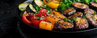 Grilled colorful bell peppers, zucchini, and eggplant slices with herbs on black plate against dark background, close up view of roasted seasonal vegetables.