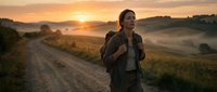 Young woman with dark hair and olive complexion wearing a backpack, standing on a rural dirt road during golden hour sunrise with misty fields and rolling hills in background.