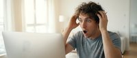 Shocked man with curly dark hair and stubble, wearing gray t-shirt, sitting at laptop with surprised expression, hand to head, in bright home office with window light.