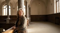 Mature woman with long gray hair and fair complexion, wearing a brown cardigan, seated in a historic stone church interior with arched ceilings and wooden pews, contemplative expression.