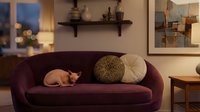 Ginger cat resting on burgundy velvet sofa in modern living room with warm lighting, decorative pillows, wooden furniture and artwork on walls.