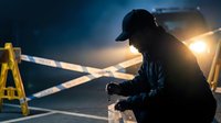Construction worker in safety gear operating equipment at dawn with bright work lights illuminating industrial job site during early morning shift.