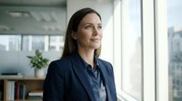 Confident female professional with long dark hair wearing navy blazer gazes out office window, modern workplace setting with cityscape view.