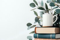 White ceramic mug on stack of vintage books with fresh eucalyptus branches against white background, creating minimal aesthetic composition.