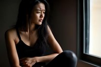 Young Asian woman in black tank top sitting alone by window with concerned expression, dark moody lighting creates emotional atmosphere for mental health concepts.