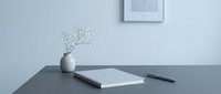 Minimalist workspace with notebook, pen, and white flower vase on gray desk, framed art on wall, clean modern home office aesthetic.