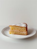 Slice of vanilla sponge cake with cream filling and powdered sugar dusting on white plate against neutral background, minimalist food photography styling.