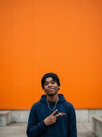 Young man, Black ethnicity, wearing dark blue hoodie and black beanie, pointing at chest against vibrant orange wall background. Suitable for youth lifestyle, streetwear, urban fashion, and casual portrait projects.