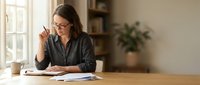 Professional businesswoman with glasses writing notes at wooden desk in bright home office with natural lighting and plants.