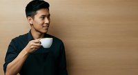 Young Asian man in black casual shirt holding white cup against beige wall, looking away thoughtfully, suitable for lifestyle and beverage marketing campaigns.