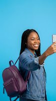 Happy African American student woman taking selfie with smartphone wearing denim jacket and backpack on blue background for social media content.
