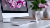 Modern workspace with computer monitor, wireless keyboard on white desk with pink cherry blossom flowers and glass of water, home office setup in soft daylight.