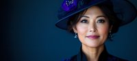 Asian woman wearing elegant dark blue hat with floral decoration and matching attire smiling confidently against deep blue background in dramatic lighting.