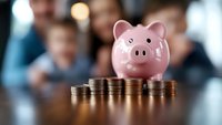 Pink ceramic piggy bank with stacks of coins arranged in ascending order, symbolizing financial growth and savings goals against blurred business meeting background.