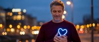 Middle-aged Caucasian man holding smartphone with glowing blue heart icon on screen during evening twilight in urban setting with bokeh city lights.