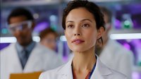 Young Hispanic female medical professional in white lab coat with confident expression in modern laboratory setting with diverse colleagues in background.