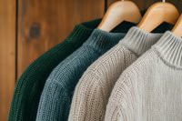 Warm knitted sweaters in neutral colors hanging on wooden hangers against rustic background. Close up view shows detailed wool texture and stitching patterns.
