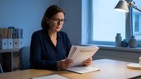 Professional middle-aged businesswoman with glasses reading documents at modern office desk with lamp, focused on paperwork analysis and review.