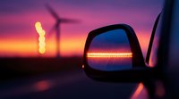 Side mirror reflection at sunset with wind turbine silhouette and vibrant purple sky. Dramatic evening landscape with renewable energy installation and sun flare.